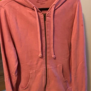 Rosa hoodie med dragkedja och huva - Mysig rosa hoodie med dragkedja framtill, huva och snörning. Tröjan har två stora fickor på magen och långa ärmar. Perfekt för en avslappnad stil och enkel att matcha med jeans eller leggings.