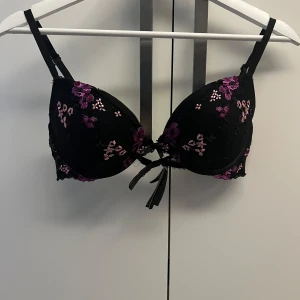 Svart blommig bh från Victoria's Secret - Supersnygg svart bh från Victoria's Secret med broderade blommor i lila och rosa. Bh:n har vadderade kupor, justerbara axelband och en liten rosett framtill. Utsidan är dekorerad med spets och meshdetaljer för en extra lyxig känsla. Storlek 75 c 