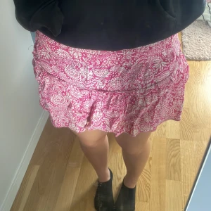 Rosa blommig volangkjol - Supersöt kort kjol i rosa och vitt med blommigt mönster och volangdetalj. Perfekt för dig som gillar en feminin och lekfull stil. Kjolen har en bekväm passform och är lätt att matcha med en enkel topp.