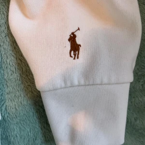 Vit hoodie från Polo Ralph Lauren - Vit hoodie från Polo Ralph Lauren med stor svart POLO-text och broderad logga på ärmen. Tröjan har huva med snörning, ribbade muddar och är tillverkad i mjuk bomull. Perfekt för chill dagar och har en klassisk, clean look.
