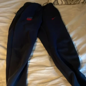 Mörkblå Nike mjukisbyxor XS - Mörkblå mjukisbyxor från Nike i storlek XS. Byxorna har dragsko i midjan, diskret Nike-logga i rött och praktiska fickor med dragkedja. Perfekta för chill eller träning, med en skön loose passform och sportig look.