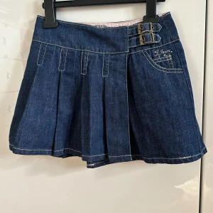 Jeanskjol med spännen från J Jeans - Mörkblå barnjeanskjol 7-9 år från J Jeans med två snygga metallspännen i midjan och dekorativ ficka fram. Kjolen har lagda veck och vit kontrastsöm, samt omlott-design framtill. 