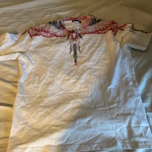 Vit Marcelo Burlon t-shirt med röd print - Säljer en vit t-shirt från Marcelo Burlon med unikt rött och svart print över axlar och hals. T-shirten är i 100% bomull och har en normal passform med korta ärmar. Perfekt för dig som vill sticka ut med en cool design.