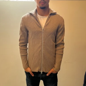 Beige zip up från Ljung by Marcus Larsson  - Säljer en stilren beige ribbad kofta från Ljung by Marcus Larsson. Koftan har hel dragkedja framtill, hög krage och är tillverkad i ett mjukt stickat material. Perfekt för lager-på-lager och passar till jeans eller chinos. NYPRIS:1699Kr MITT PRIS:499Kr✅