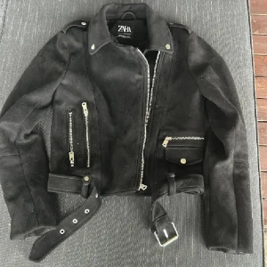 Svart mocka bikerjacka från Zara - Säljer en svart bikerjacka i mocka från Zara. Jackan har silvriga dragkedjor, bälte i midjan och flera fickor med dragkedja och knappdetaljer. Klassisk krage och långärmad modell som ger en cool och edgy look. Använd gärna köp nu