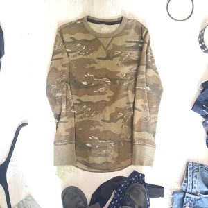 Camouflage tröja från Hollister - Snygg långärmad tröja från Hollister i klassiskt camouflage-mönster. Tröjan har rund halsringning och ribbade muddar vid ärmslut och nederkant. Perfekt för dig som gillar streetstyle och vill sticka ut lite extra.