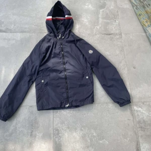 Mörkblå vindjacka från Moncler - Snygg mörkblå vindjacka från Moncler med huva och dragkedja. Jackan har Moncler-logga på ärmen och röda samt vita ränder på huvan. Två fickor framtill med knappar och elastiska muddar vid ärmslut. Perfekt för blåsiga dagar.
