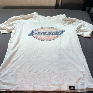 Vit Dickies t-shirt med stor logga - Vit t-shirt från Dickies i storlek XS med stor klassisk Dickies-logga i blått och rött på bröstet. T-shirten är i mjuk bomull och har en normal passform med korta ärmar. Perfekt för en avslappnad streetwear-look.