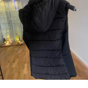 Svart dunväst från Zara med huva - Svart quiltad dunväst från Zara med dragkedja och stor huva. Västen är lång och har en clean, stilren look med horisontella sömmar. Perfekt för lager-på-lager och funkar till många outfits.