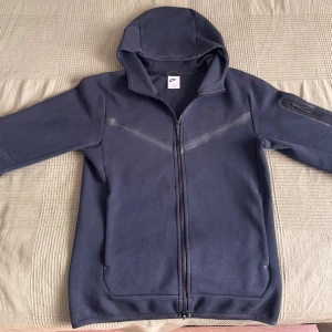 Svart Nike tech  med dragkedja XS - Svart hoodie från Nike i storlek XS med hel dragkedja och huva. Snygga detaljer med diskreta svarta ränder över bröstet och ficka med dragkedja på ärmen. Perfekt för dig som gillar sportig och clean stil. Bra skick. Igensytt hål på baksidan, men man ser inte det så tydligt bara om man fokuserar på det.
