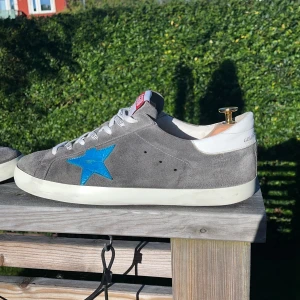 Golden Goose Superstar Storlek 44 - Golden Goose Superstar sneakers i grå mocka med vit läderdetalj på hälen och vit sula. Snygg blå stjärna i alligator skin på sidan och vita snören med GGDB-tryck. Platt svart sula med fiskbensmönster. Röd GGDB-logga på tungan. Har många fler bilder som inte fick plats det är bara att fråga. KVITTO Finns bara att fråga om bild. Köpta på farfetch För ~5000 kr. Priset går att diskutera vid snabb affär!