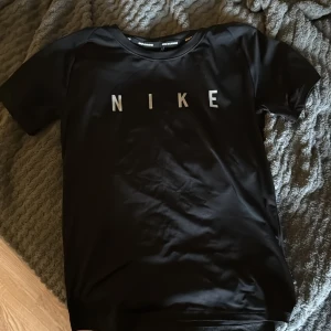 Svart Nike Dri-Fit t-shirt XS - Svart t-shirt från Nike med Dri-Fit-teknologi och stor logga framtill. Perfekt för träning, tillverkad i ett lätt och snabbtorkande syntetmaterial. Klassisk passform med rund hals och korta ärmar. Snygg och sportig stil för dig som gillar att röra på dig.