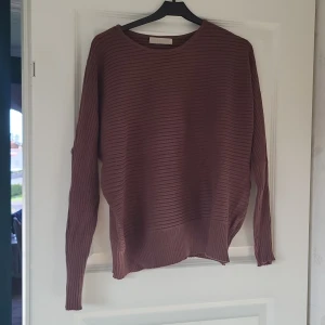 Ribbad brun tröja och kjol från Voyelles - Ribbad brun långärmad tröja från Voyelles med avslappnad passform och rund halsringning. Tröjan är stickad i ett mjukt material och har diskreta horisontella ränder för en stilren look. Perfekt för lager-på-lager under höst och vinter. Ingår en kjol av samma kvalitet.