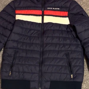 Mörkblå pufferjacka Race Marine - Mörkblå pufferjacka från Race Marine med horisontella röda och vita ränder över bröstet. Jackan har dragkedja framtill, två fickor med dragkedja och ribbade muddar. Perfekt för kyliga dagar och snygg till jeans.
