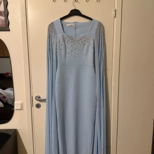 Ljusblå långklänning  - Superfin ljusblå långklänning från Rojda med glittriga pärldetaljer över byst och axlar. Klänningen har långa, svepande ärmar och en elegant slits bak. Perfekt för dig som vill sticka ut med en unik och glamorös look.