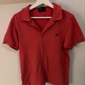 Röd Fred Perry pikétröja - Säljer en klassisk röd pikétröja från Fred Perry med broderad logga på bröstet. Tröjan har korta ärmar, krage och är gjord i mjuk bomull. Perfekt för en stilren och sportig look. Är använd men i bra skick.