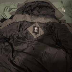 Svart dunjacka från Canada Goose - Säljer en svart dunjacka från Canada Goose med klassisk patch på ärmen. Jackan har huva, dragkedja och knappar framtill samt breda, quiltade sektioner för extra värme. Perfekt för kalla vinterdagar och riktigt snygg streetstil.