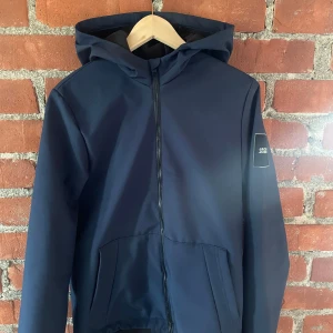 Mörkblå vindjacka från Jack & Jones - Snygg mörkblå vindjacka från Jack & Jones med huva och hel dragkedja framtill. Jackan har två fickor och en stilren patch på ärmen. Perfekt för dig som vill ha en enkel och clean look.