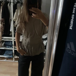 Beige blus med volang och smock - Säljer en grön blus med korta, vida ärmar och smockdetaljer i midjan som ger en snygg form. Blusen har volang nertill och är tillverkad i ett lätt och luftigt material, perfekt för varmare dagar. Enkel och stilren design som passar till mycket.