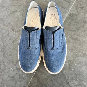 Blåa jeansloafers från Tod’s - Snygga loafers från Tod's i blått jeanstyg med vita sulor och flätad detalj runt kanten. Skorna har en stilren design utan snörning och inslag av svart skinn vid öppningen. Perfekta för dig som gillar en avslappnad men trendig look.