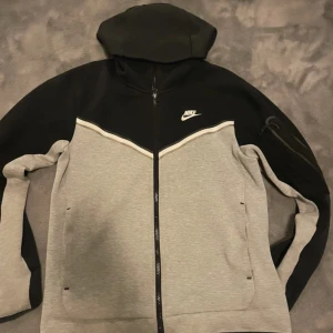 Nike Tech Fleece hoodie svart/grå - Nike Tech Fleece hoodie i svart och ljusgrått med hel dragkedja och huva. Jackan har en sportig look med vit Nike-logga på bröstet, fickor framtill och en extra ficka med dragkedja på ärmen. Perfekt för chill eller träning.