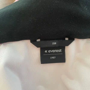 Rosa regnjacka från Everest med huva - Säljer en rosa regnjacka från Everest i storlek 158. Jackan har huva, vita tryckknappar och praktiska fickor framtill. Perfekt för regniga dagar och har en stilren look med reflexdetalj på ryggen. Materialet är syntet och jackan är helfärgad.