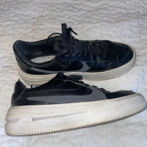 Nike Air Force 1 svarta sneakers - Svarta Nike Air Force 1 sneakers med grå swoosh och vit, tjock sula. Klassisk modell med snörning och rund tå. Ovandel i skinn och tydlig AIR-detalj på sulan. Perfekta för dig som gillar streetstyle och vill ha en ikonisk sneaker i garderoben. Inner sula saknas. 