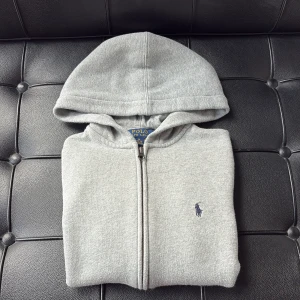 Grå hoodie från Polo Ralph Lauren - Grå hoodie från Polo Ralph Lauren med dragkedja framtill och klassisk huva. Tröjan har broderad logga på bröstet och är tillverkad i mjuk bomullsblandning. Perfekt tröja att ha oavsett tillfälle. Tveka inte på att skriva om ni har några frågor eller funderingar!