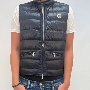 Svart dunväst från Moncler - Snygg svart dunväst från Moncler med klassisk quiltad design och hög krage. Västen har två dragkedjefickor framtill och stängs med dragkedja. Ikonisk Moncler-logga på bröstet. Perfekt att slänga över en hoodie eller t-shirt.
