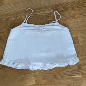 Vit och rosa randig linnetopp med volangkant - Supersöt vit och rosa randig linnetopp med tunna axelband och spetsliknande detaljer vid toppen. En jättesöt volang vid botten som lägger till en extra gullig touch. Säljer för att jag köpte i en för liten storlek💞