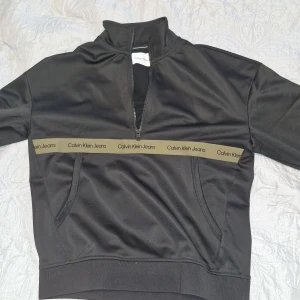 Svart Calvin Klein half zip tröja XS - Svart tröja från Calvin Klein Jeans med halv dragkedja och hög krage. Snygg detalj med olivgrön rand och logga över bröstet. Praktiska fickor framtill och sportig look. Perfekt för dig som gillar stilrena och bekväma plagg.