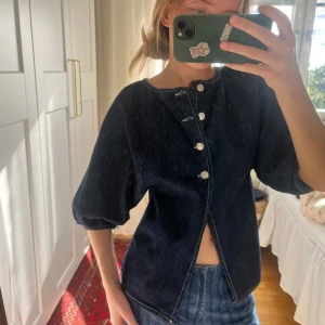 Mörkblå jeansblus med puffärm - Säljer en mörkblå jeansblus med trekvarts puffärmar och snygga silvriga knappar framtill. Blusen har en rak passform och är tillverkad i jeansmaterial med synliga sömmar. Perfekt för dig som gillar en cool och avslappnad stil.