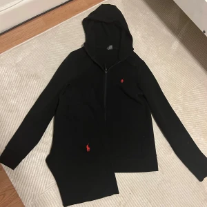 Svart hoodie från Ralph Lauren - Svart hoodie från Ralph Lauren med dragkedja och huva. Klassisk röd broderad logga på bröstet och ficka framtill. Perfekt för en chill och stilren look. Långa ärmar och mjukt material som känns skönt mot huden.