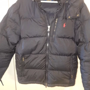 Svart pufferjacka från Polo Ralph Lauren - Säljer en svart pufferjacka från Polo Ralph Lauren med huva och dragkedjor. Jackan har en klassisk röd logga på bröstet, flera fickor med dragkedja och är vadderad för extra värme. Perfekt för kalla dagar och riktigt snygg streetstil.Jacka har ett litet hål som har sytt ihop.