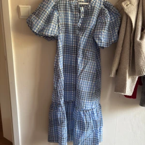 Blårutig klänning med puffärm från Vila - Supersöt klänning från Vila i blått och vitt rutigt mönster. Modellen har puffiga korta ärmar, knappar framtill och volang nertill. Perfekt för dig som gillar en luftig och trendig look. Tillverkad i lätt bomullsmaterial.