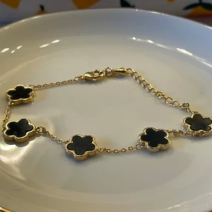 Guldfärgat armband med svarta blommor - Snyggt armband med tunna guldfärgade länkar och fem svarta blommor i klöverform. Blommorna har en elegant inramning i guldton och armbandet stängs med ett klassiskt lås. Perfekt accessoar för att lyfta din outfit.