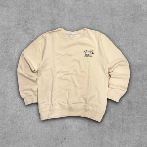 Beige sweatshirt med tryck Fast Food - Beige sweatshirt med rund hals och trycket 'Fast Food' på bröstet. Tröjan har långa ärmar, ribbade muddar och avslappnad passform. Perfekt för en chill och avslappnad stil.