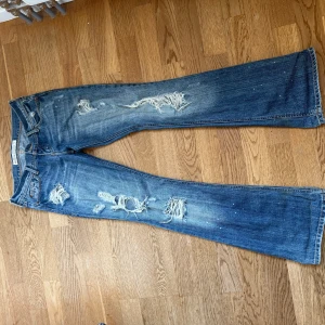 Blå bootcut jeans med slitningar - Snygga blå jeans från Miss Sixty med bootcut-modell och slitna detaljer framtill. Jeansen har coola broderade bakfickor och små vita färgstänk för en extra edge. Klassisk femficksdesign och låg midja. Perfekta för dig som gillar en avslappnad och trendig stil.