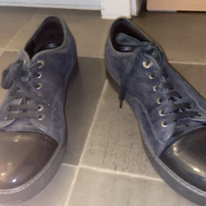 Grå mockasneakers från Lanvin - Snygga sneakers från Lanvin i mörkgrå mocka med matchande grå snörning och gummitå. Skorna har en platt sula och rund tå, vilket ger en clean och stilren look. Perfekta för dig som gillar exklusiva och minimalistiska sneakers med premiumkänsla.
