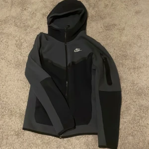 Nike svart och grå vindjacka XS - Nike vindjacka i svart och mörkgrått med huva och hel dragkedja. Jackan har en sportig look, diskret Nike-logga på bröstet och praktisk ficka med dragkedja på ärmen. Tillverkad i mjukt syntetmaterial, perfekt för aktiva dagar.