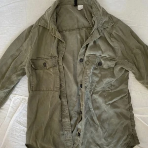 Olivgrön overshirt från Divided H&M - Olivgrön overshirt från Divided H&M i bomull. Skjortan har två bröstfickor med knappar, klassisk krage och knäppning framtill. Perfekt att bära öppen eller stängd för en avslappnad look.
