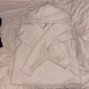 Vit hoodie från Peak Performance M - Säljer en vit hoodie från Peak Performance i storlek M. Tröjan har ett snyggt stickat kabelmönster, huva och ribbade muddar. Perfekt för dig som gillar en clean och mysig stil med sportig touch.
