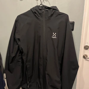 Svart vindjacka från Haglöfs med huva - Svart vindjacka från Haglöfs i storlek M. Jackan har hel dragkedja, justerbar huva och meshfoder på insidan. Perfekt för blåsiga dagar och outdoor-aktiviteter. Snygg minimalistisk design med Haglöfs-logga på bröstet. 