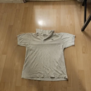 Beige pikétröja från Lacoste - Klassisk beige pikétröja från Lacoste med ikoniska krokodilloggan på bröstet. Tröjan har korta ärmar, krage och knäppning framtill. Tillverkad i bomull för en skön och luftig känsla. Perfekt för dig som gillar stilren och tidlös design.