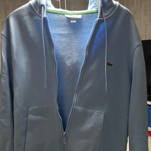 Babyblå hoodie från Lacoste - Säljer en babyblå kofta från Lacoste med dragkedja och huva. Tröjan har klassisk krokodil-logga på bröstet, två fickor framtill och justerbara snören i huvan. Perfekt för en chill och sportig stil. Köpt på Zalando för 1800kr, sparsamt använd i princip ny.