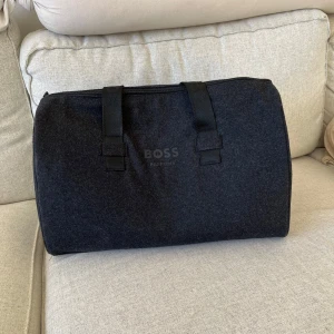 Svart/mörkgrå weekendbag från BOSS Parfums - Stilren svart weekendbag från BOSS Parfums med broderad logga på handtagen. Väskan är tillverkad i filtliknande syntetmaterial (70% polyester & 30% ull) med slät insida och har dragkedja upptill. Perfekt storlek för en kort resa eller gymmet. Ca 50cm bred, 22cm djup & 30cm hög