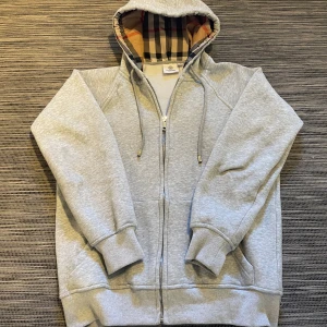 Grå hoodie från Burberry med dragkedja - Säljer en snygg hoodie från Burberry! Hoodien är köpt på NK i Göteborg när jag var där för 3799kr! Säljes för den inte passar längre. Digitalt kvitto finns😊 Hoodien är i mjukt material på insidan vilket gör den perfekt till vintern!❄️ Den har inga defekter. Om ni har några frågor så är de bara att höra av er!