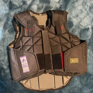 Svart säkerhetsväst från Globus Sport - Svart säkerhetsväst från Globus Sport i barnstorlek large. Västen har quiltad design med triangulära mönster och breda axelband. Kardborreknäppning framtill och på sidorna för enkel justering. Märkt med BETA Level 3 för extra skydd vid ridning. Aldrig varit med om något fall