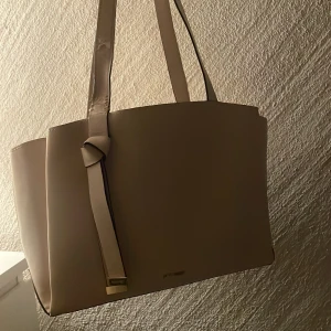 Beige axelväska från Nike west - Stilren beige axelväska i slät skinnimitation. Väskan har raka linjer, långa axelband och en snygg knutdetalj framtill med metallfäste. Perfekt storlek för att få plats med allt du behöver till vardagen.