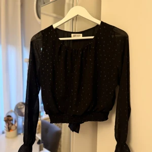 Svart blus med knyt baktill från H&M - Svart blus från H&M med små diskreta glittriga prickar. Blusen har knytning baktill och är tunn samt luftig.